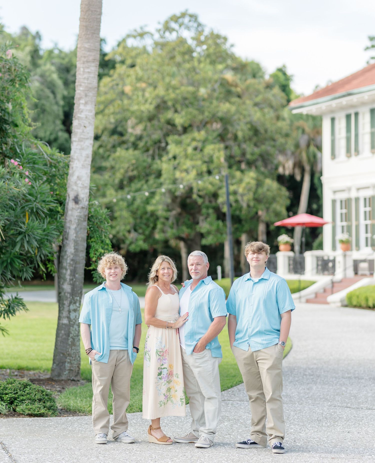 A group in coordinating pastel blue and beige outfits poses casually on a palm-lined path near a white mansion.