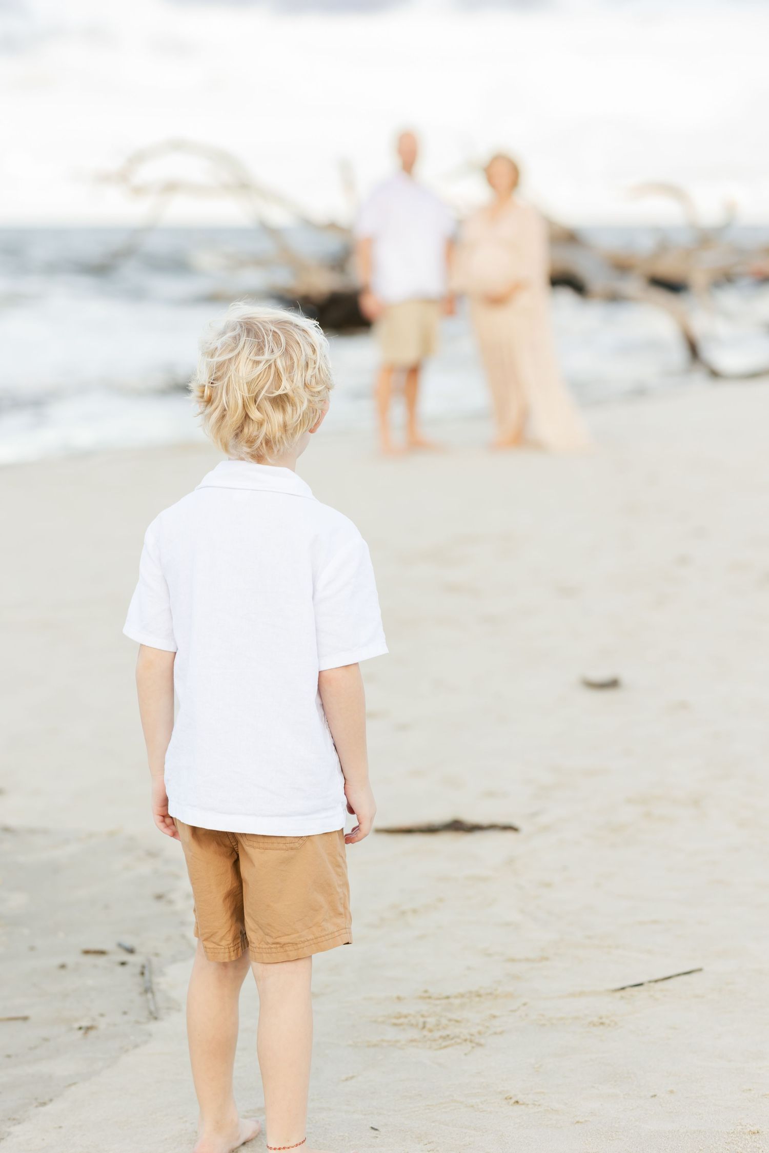 Child in white shirt watches two figures from behind on a beach with driftwood.