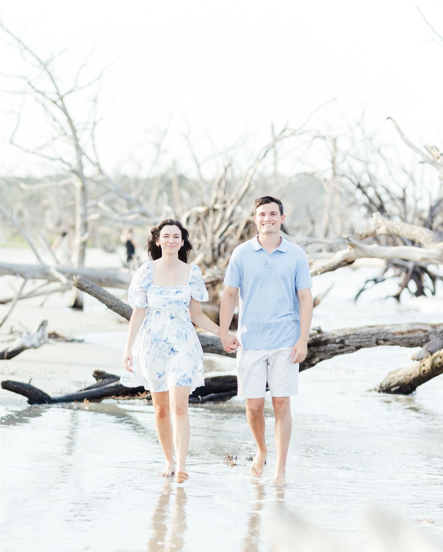 Couple walks through shallow ocean water near driftwood on a serene white sand beach.