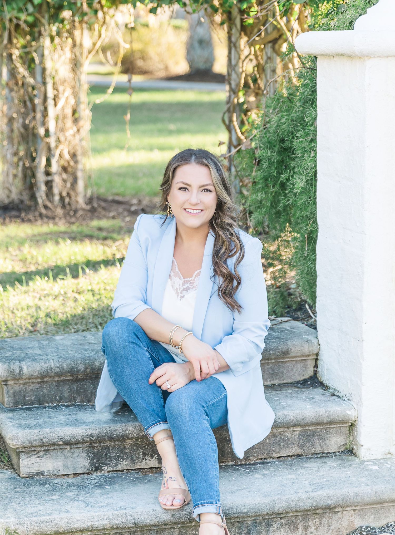 Someone in a light blue blazer and jeans sits casually on white stone steps in a sunny outdoor setting.