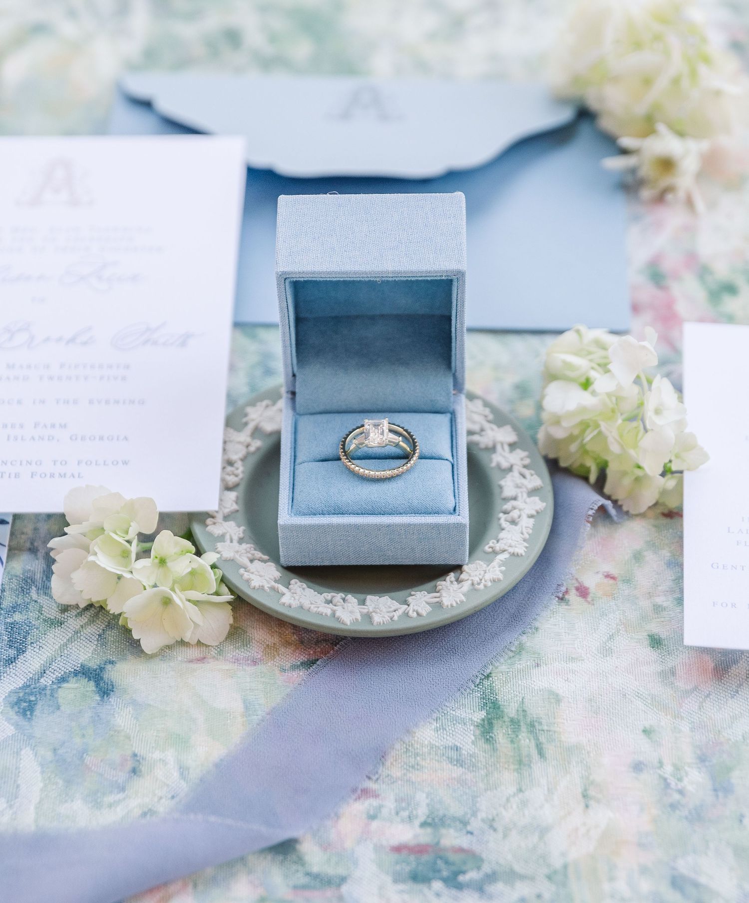 Blue velvet ring box surrounded by delicate white flowers on a textured marble surface.