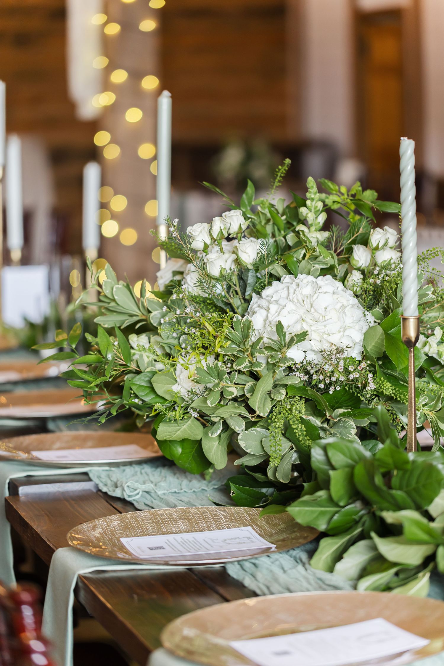 Elegant wedding table setting with white floral centerpiece and greenery garland against warm bokeh lights.