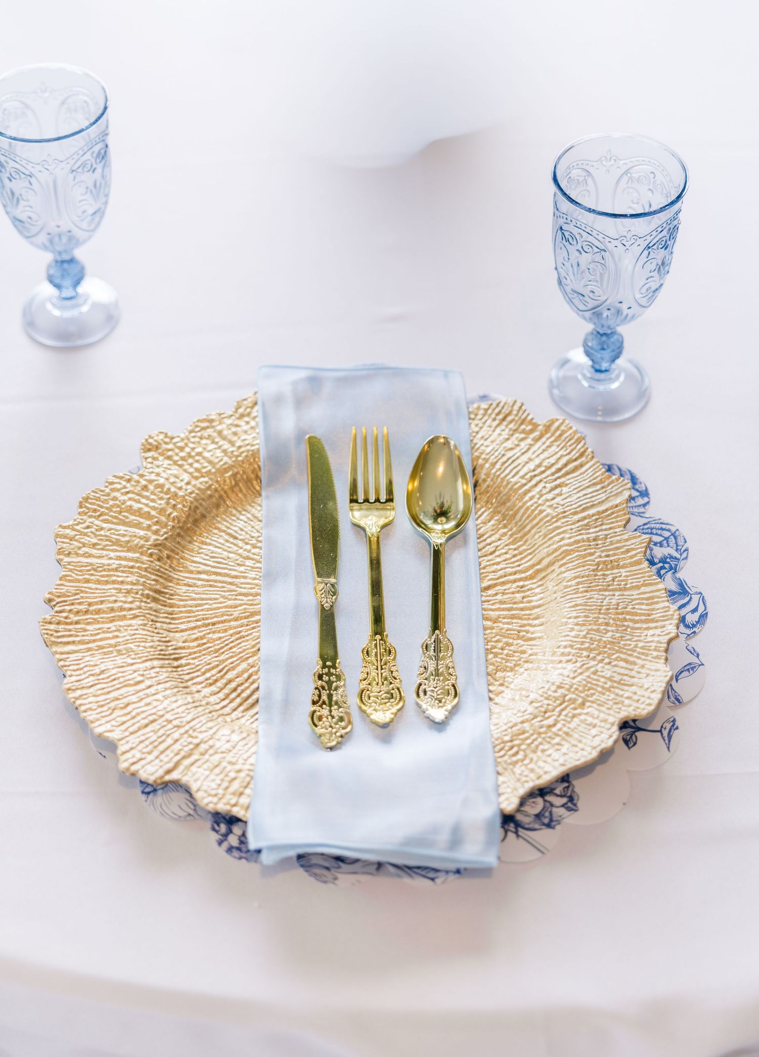 Elegant wedding place setting with gold charger plate, gold cutlery, light blue napkin, and blue crystal goblets.
