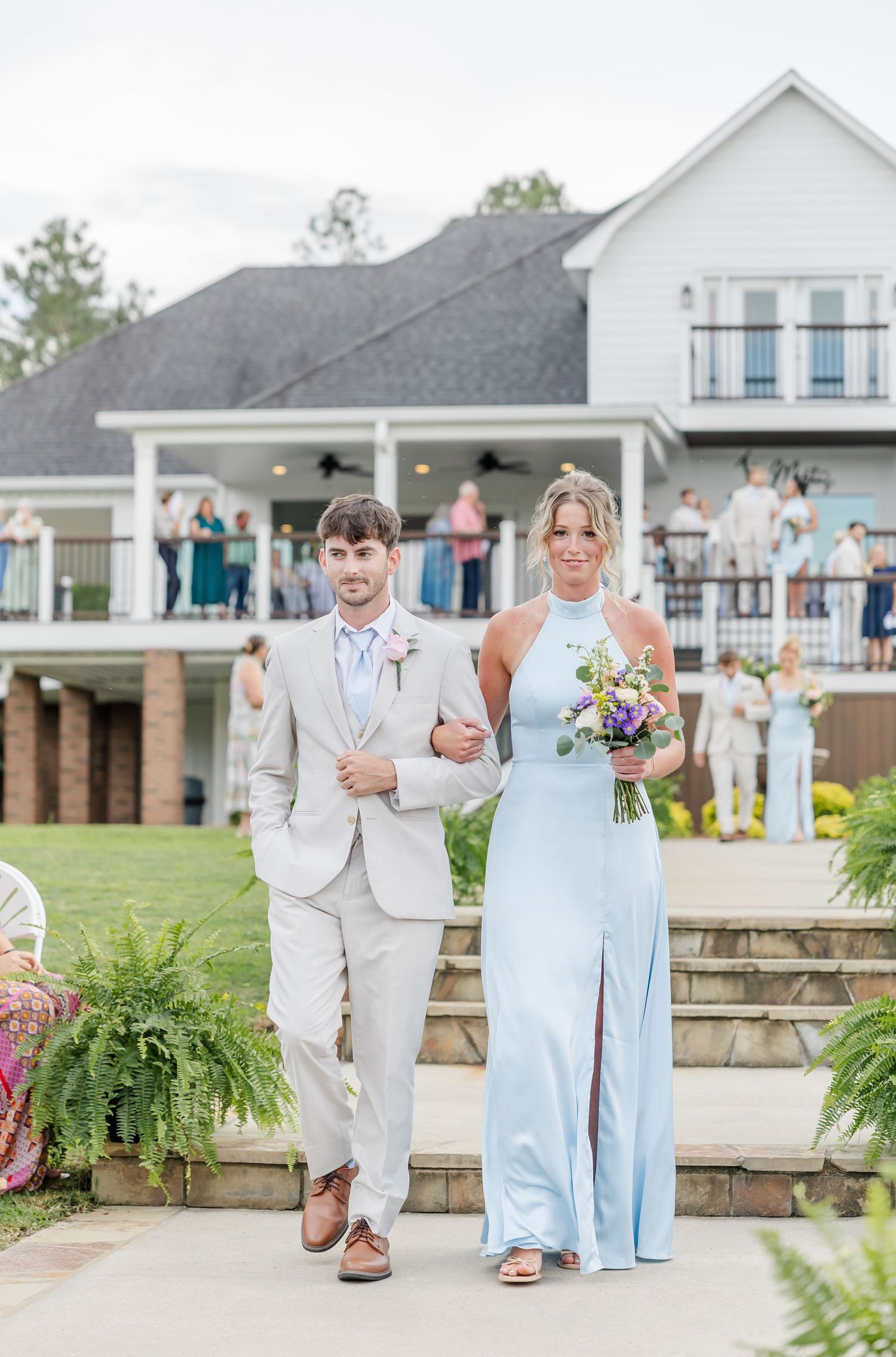 Groomsman in tan suit escorts bridesmaid in light blue gown down outdoor wedding ceremony aisle.