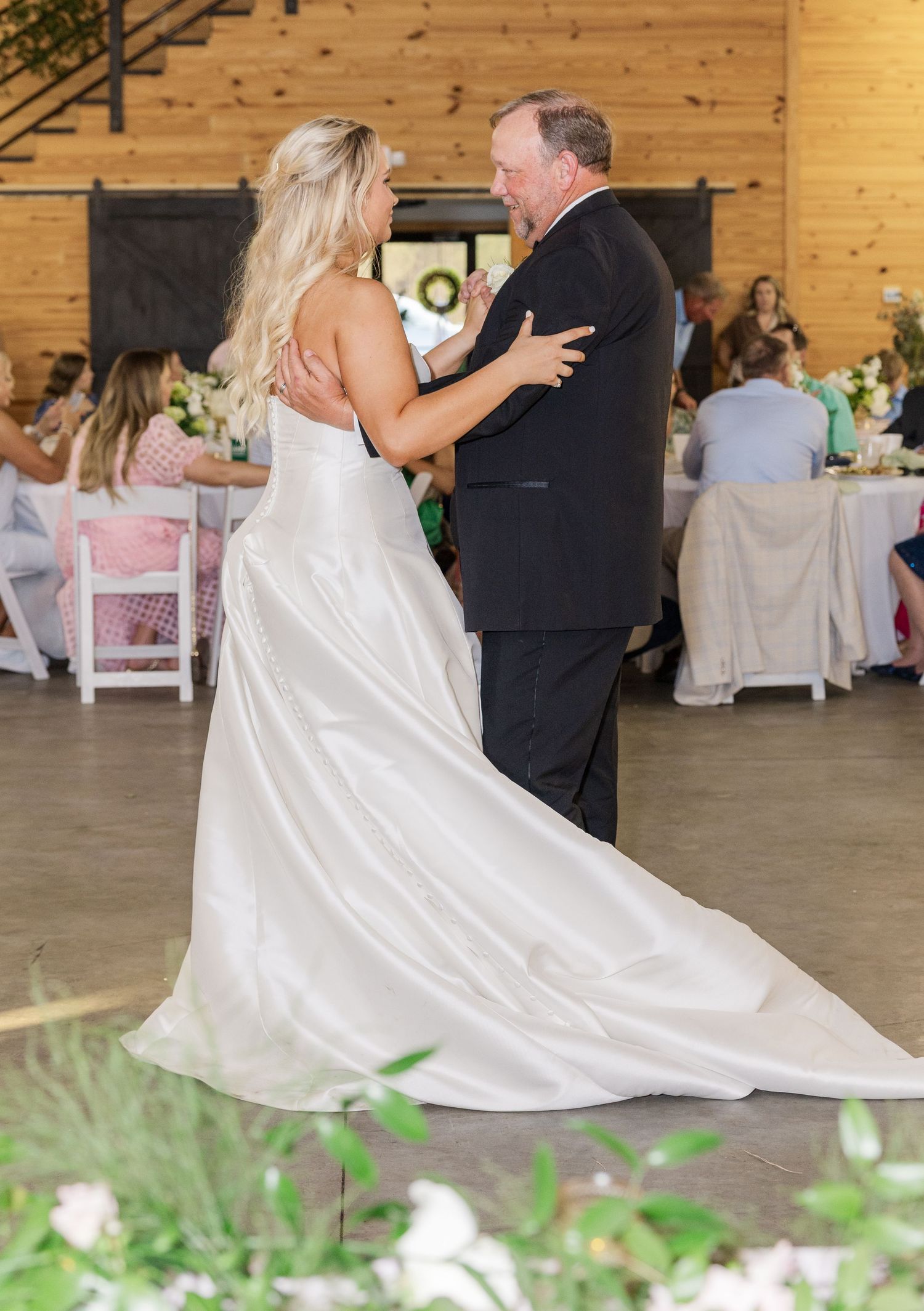 Bride in elegant white gown shares father-daughter dance inside rustic barn wedding reception venue.