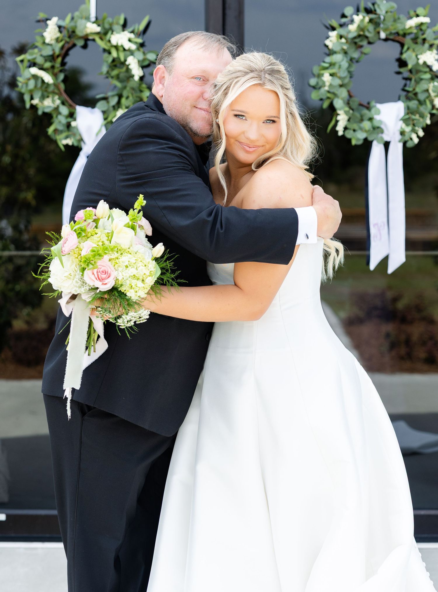 Bride in white off-shoulder gown holds pink bouquet while embracing a man in black suit outdoors.