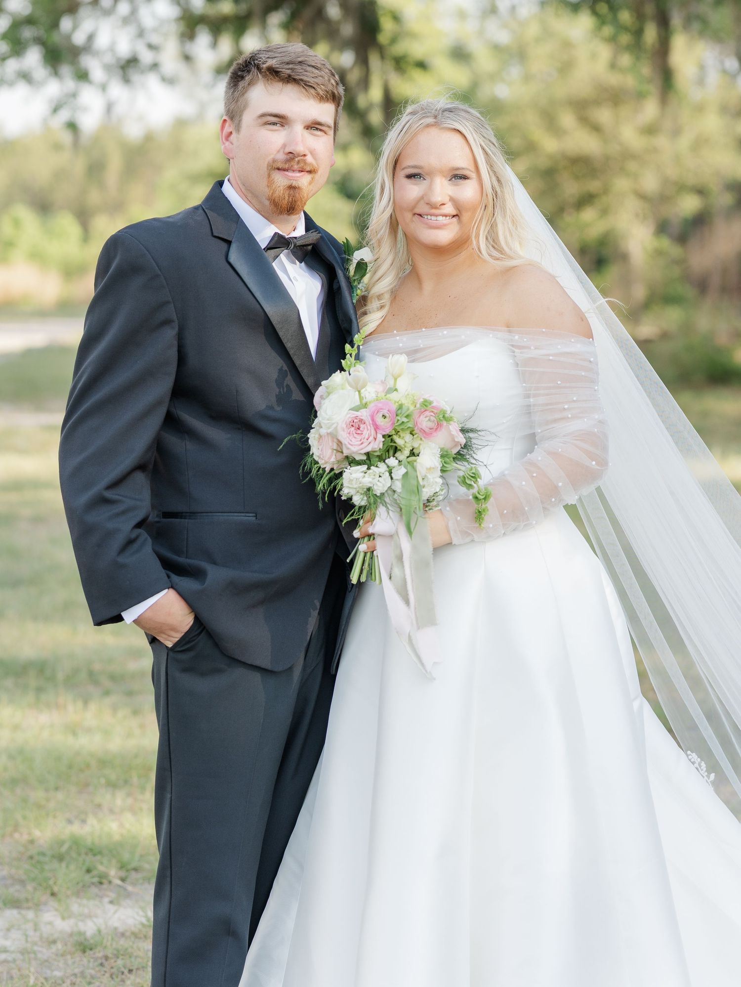 Smiling bride and groom portrait outdoors, bride wearing off-shoulder gown with veil and pink bouquet.
