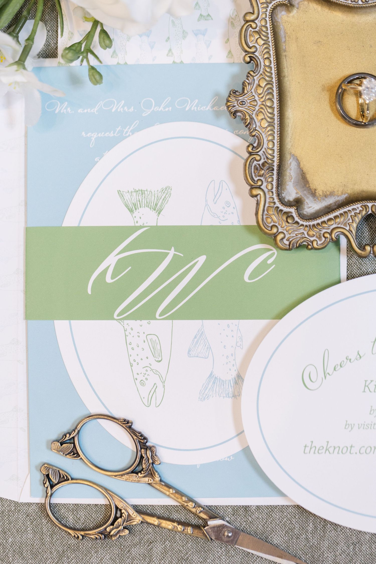 Elegant wedding invitation suite with green monogram band, gold scissors, and floral details on a light blue design.