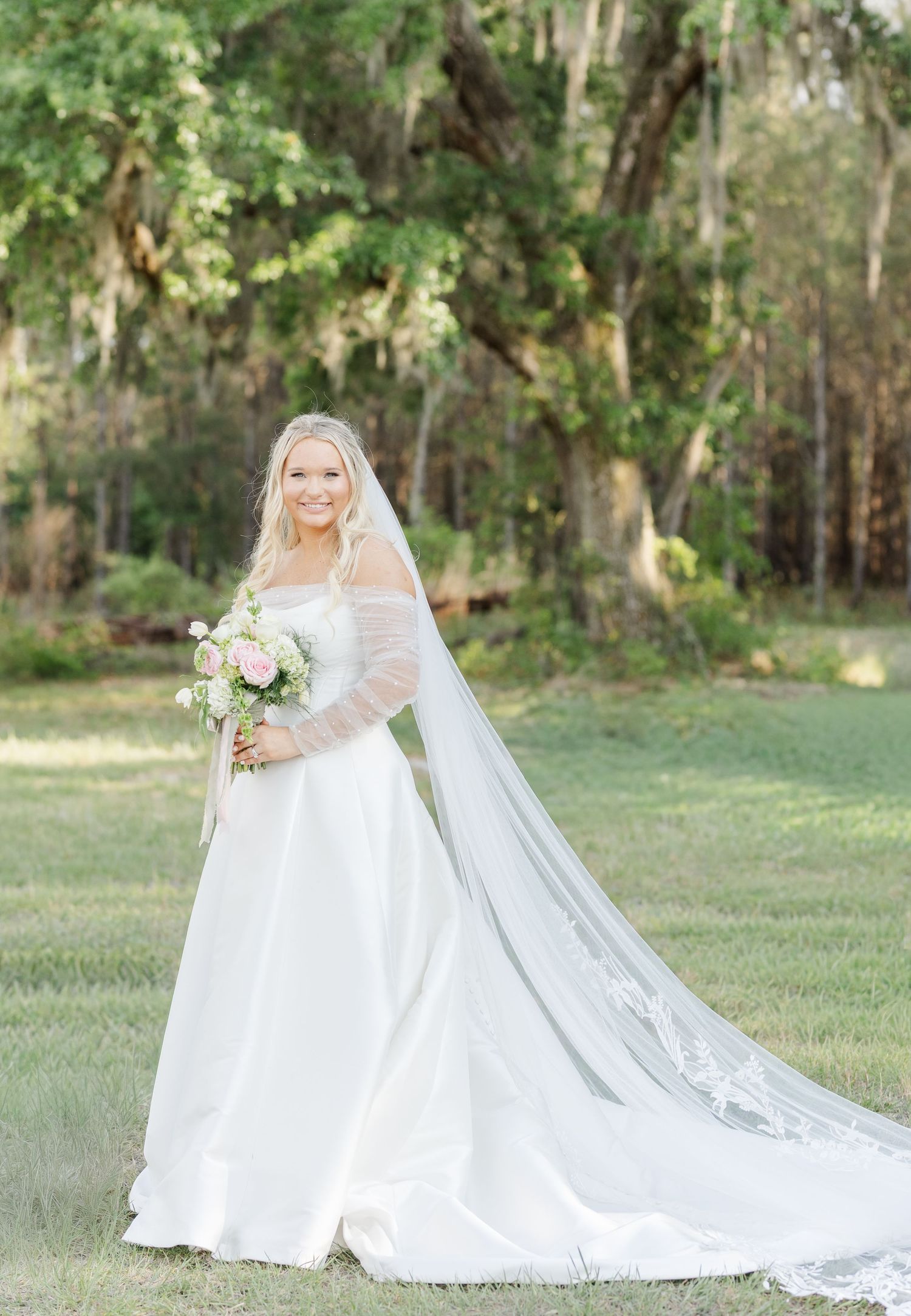 Smiling bride in white gown with long lace veil holding pink bouquet outdoors among trees.