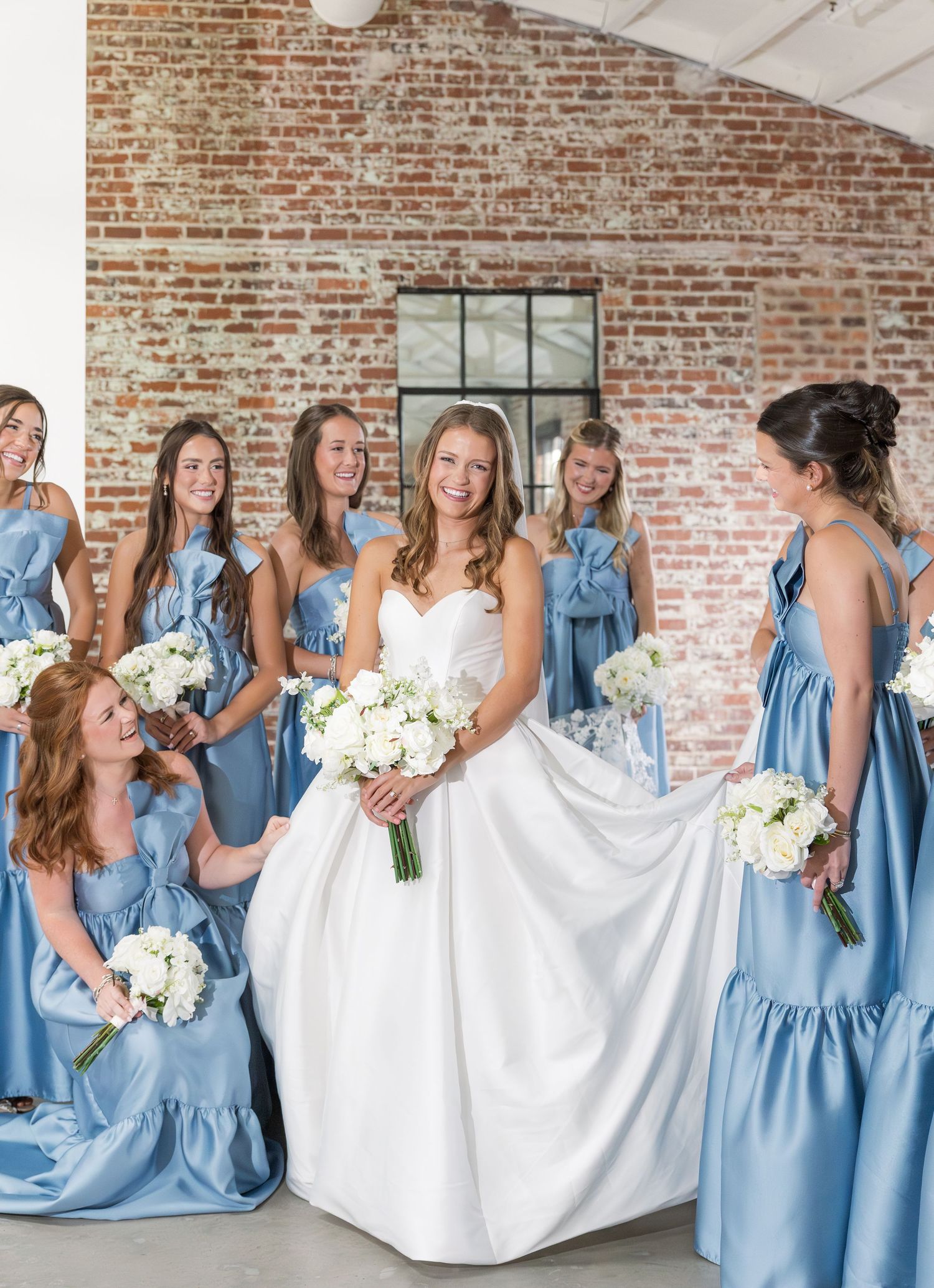 Smiling bride in white gown holds white bouquet surrounded by bridesmaids in dusty blue dresses.
