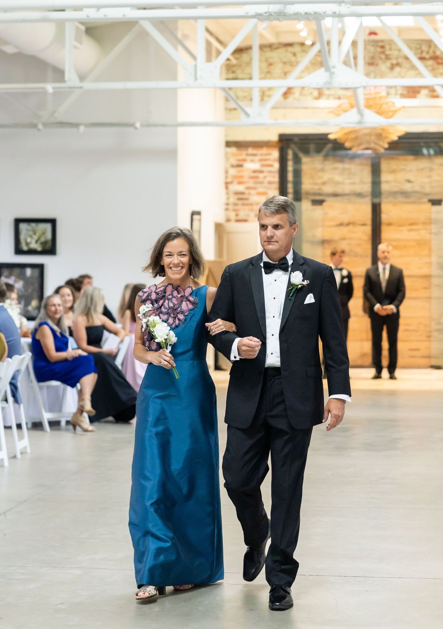 Elegant couple walks down wedding aisle, woman in teal gown holding bouquet, man in black tuxedo.