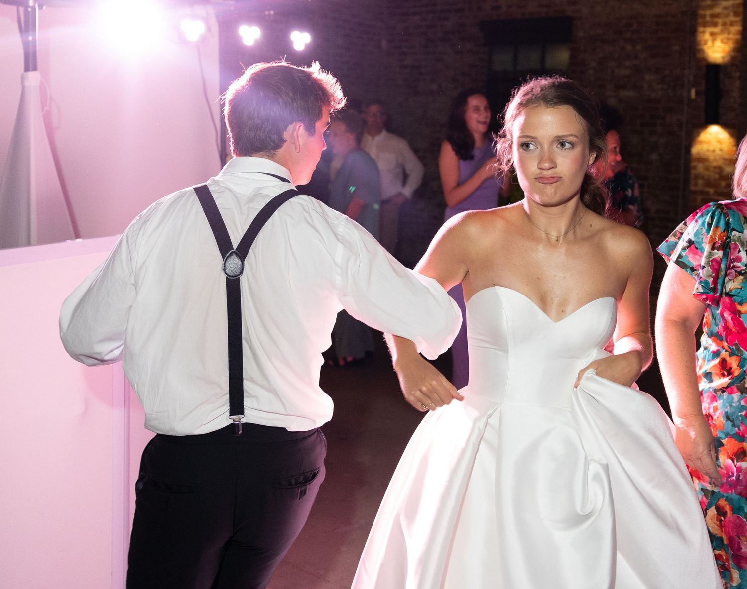 Bride in strapless gown dancing with groom in suspenders under pink lights at wedding.