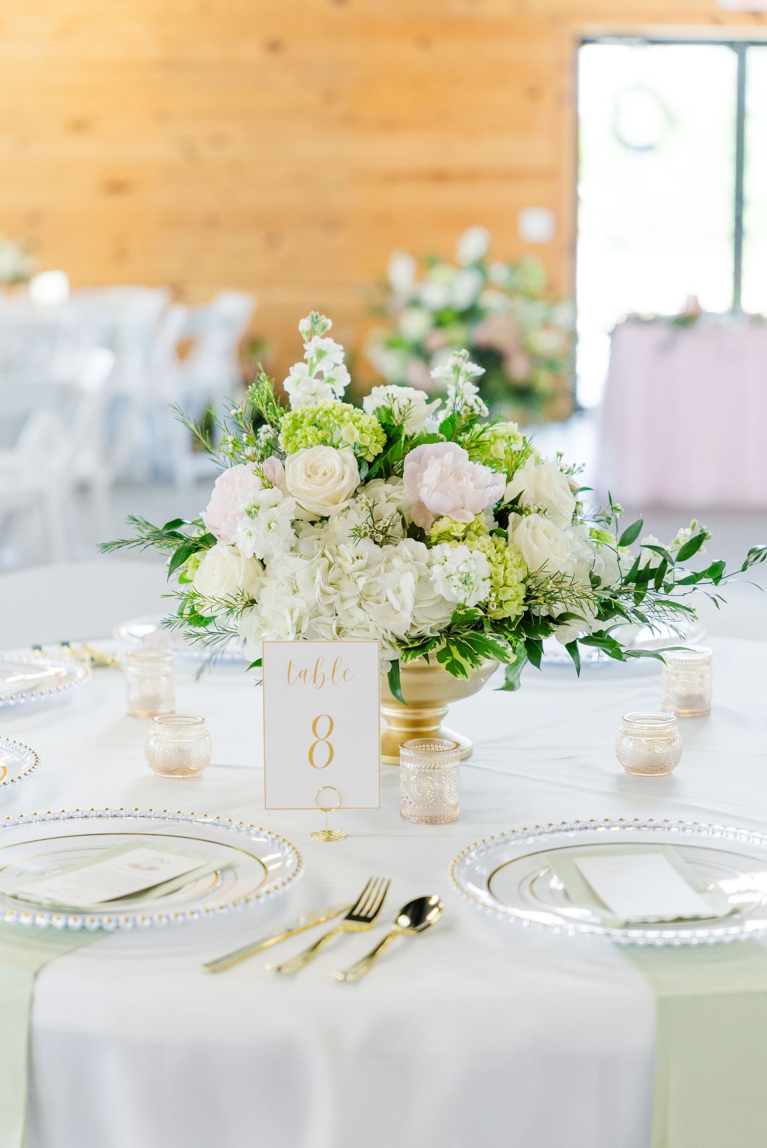 Elegant wedding table setting with gold flatware, beaded chargers, and a lush white and green floral centerpiece on table 8.