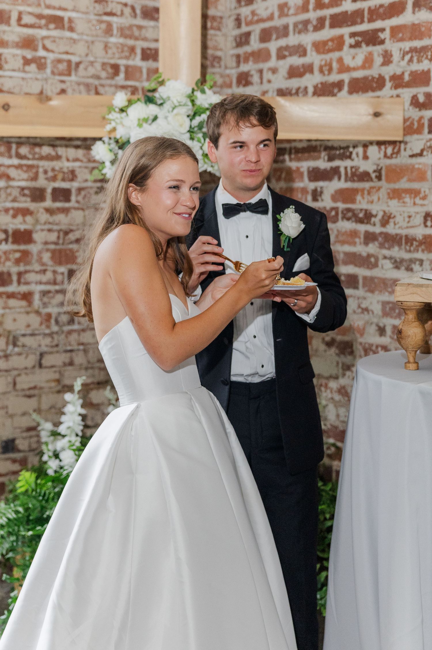Smiling bride and groom cut wedding cake together near brick wall with white floral decorations.