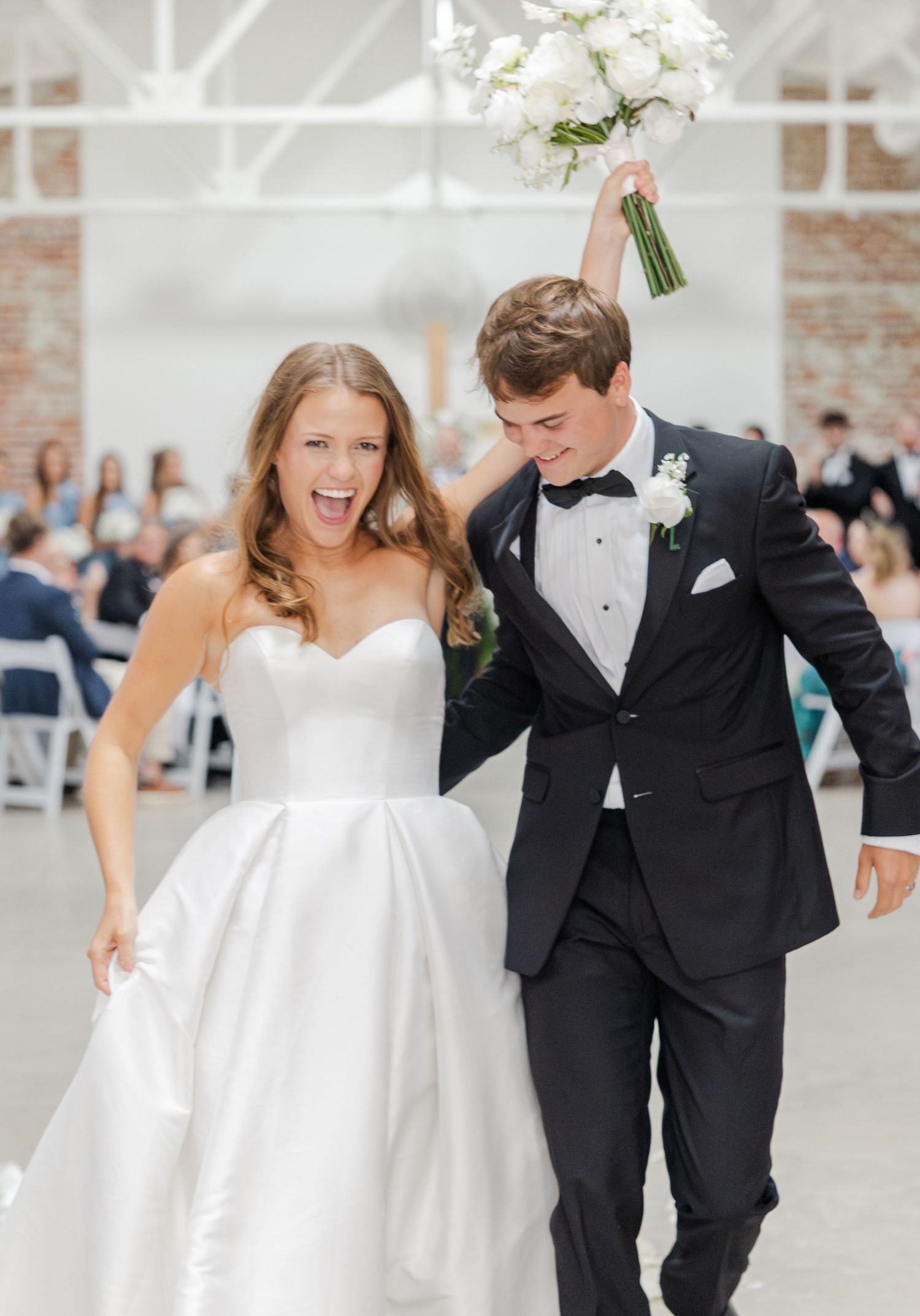 Joyful newlyweds celebrating after ceremony, bride in white gown, groom in black tuxedo, bouquet raised.