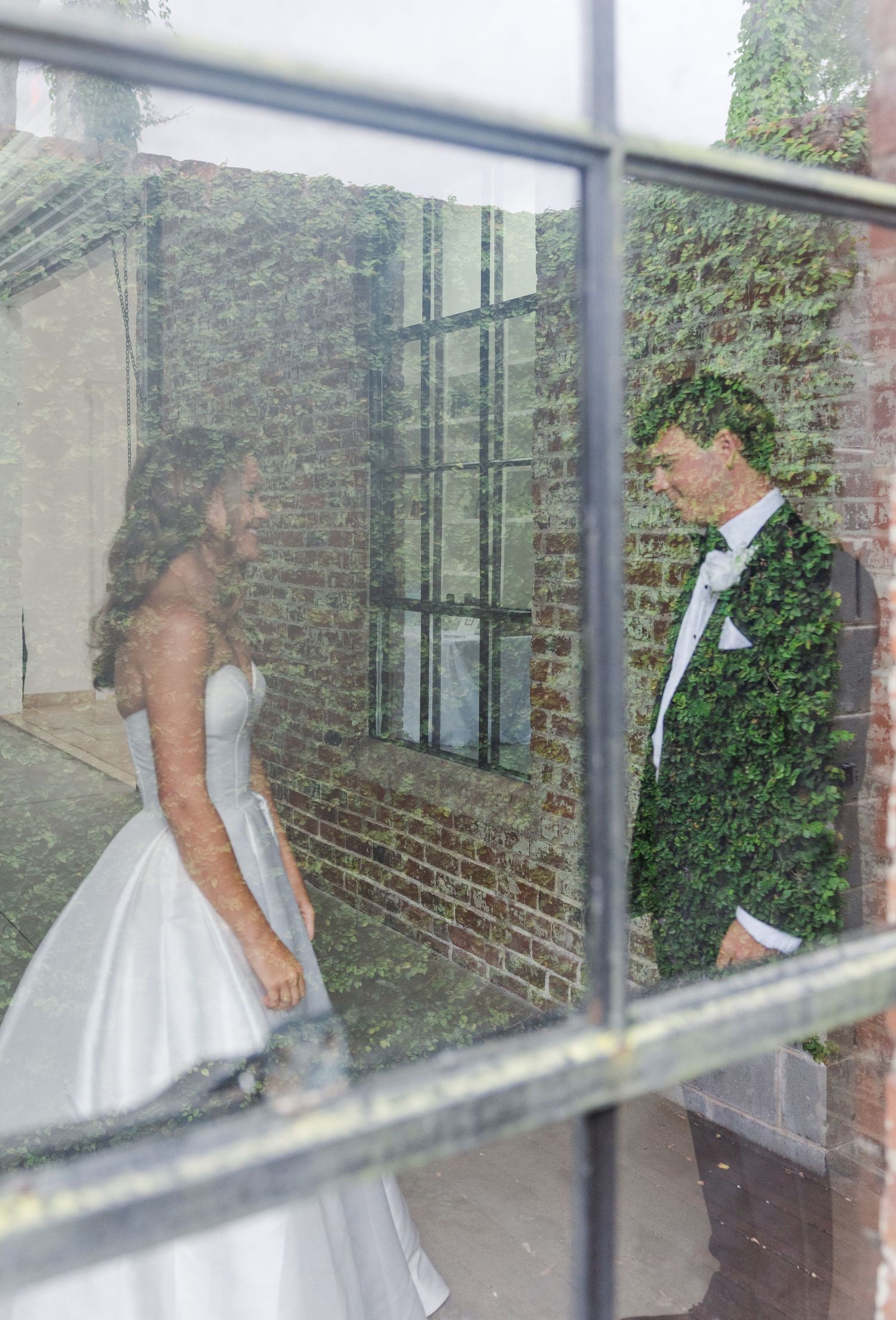 Bride and groom seen through glass window panes with ivy-covered brick wall backdrop.