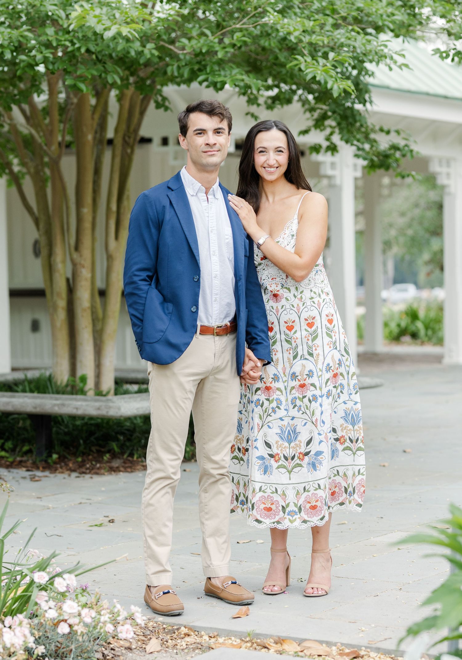 A couple poses outdoors, man in navy blazer and khakis, woman in floral midi dress, near a white building.