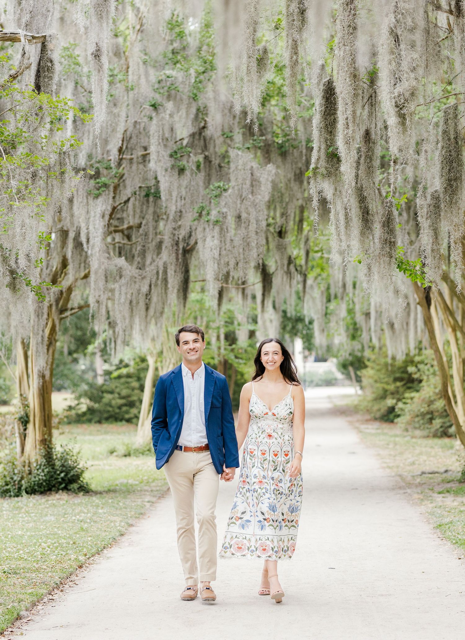 Couple walking hand-in-hand on a tree-lined path draped with Spanish moss, man in blue blazer, woman in floral dress.