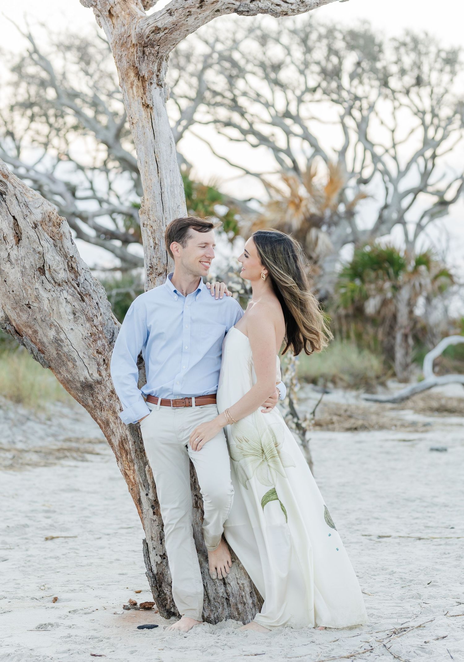 Couple sharing a romantic moment leaning against a driftwood tree on a sandy beach during golden hour.