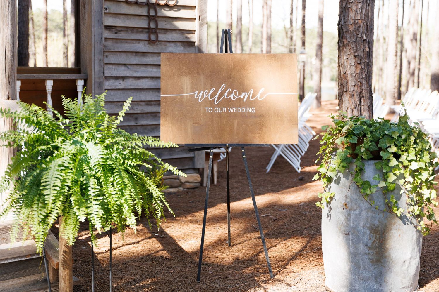 Wooden welcome wedding sign on easel flanked by lush ferns and greenery outside rustic barn venue.