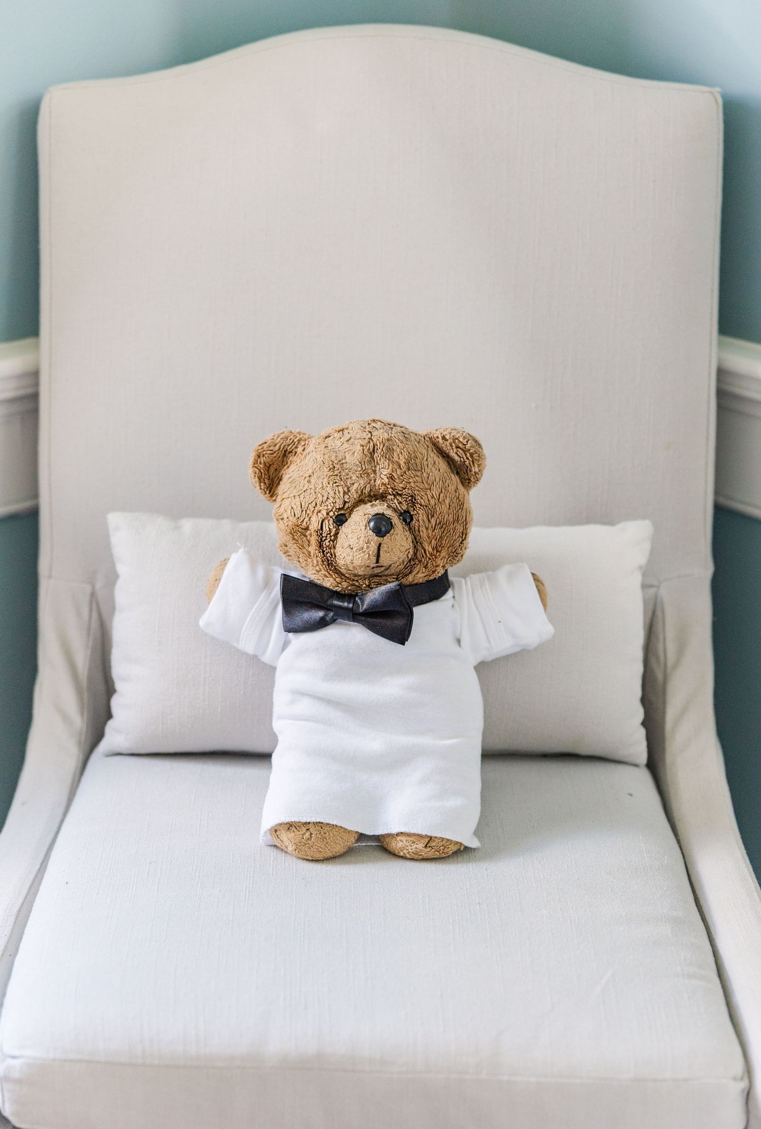 Teddy bear dressed in white tuxedo shirt and black bow tie seated on white chair.