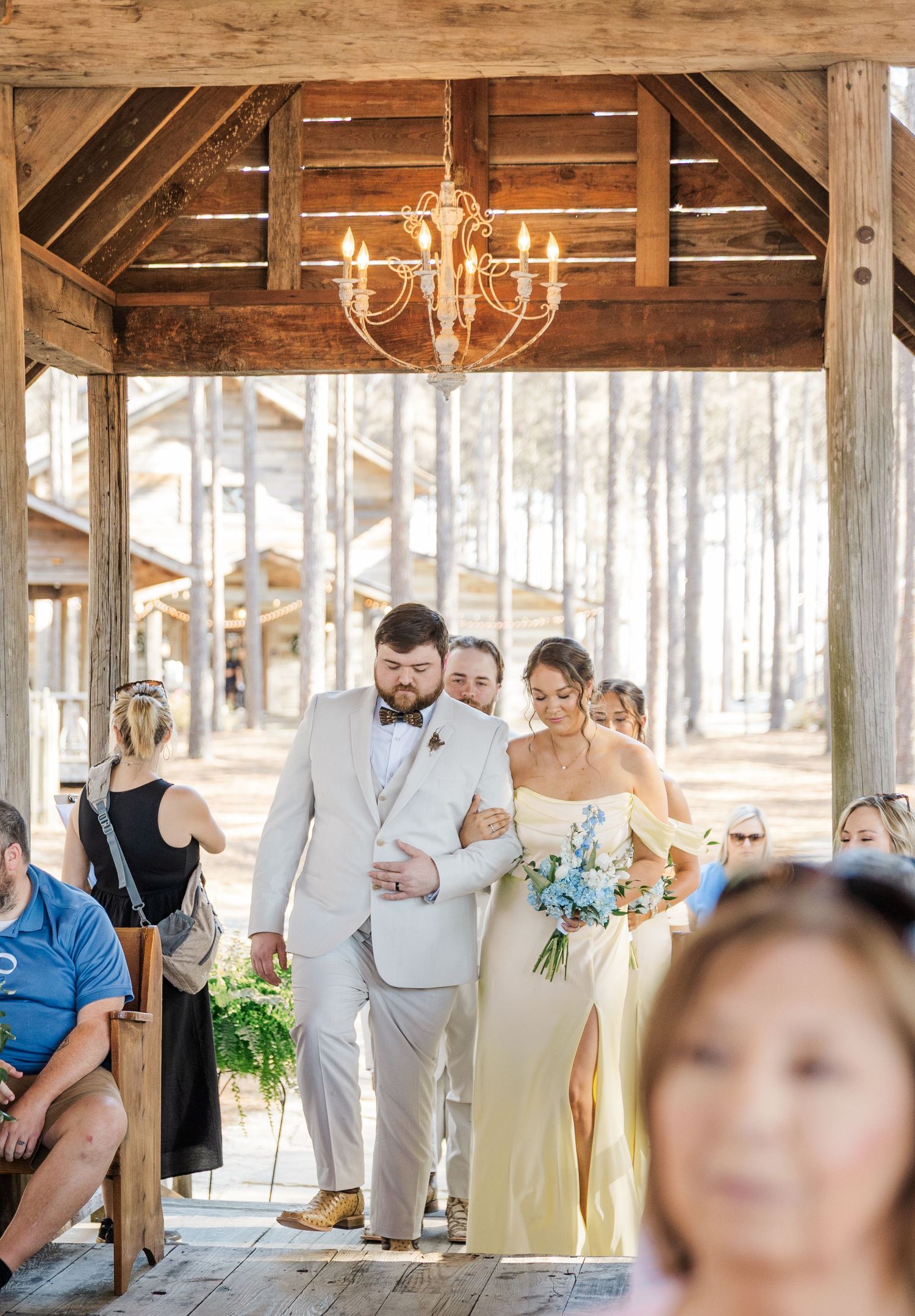 Groom in white suit escorts bridesmaid in yellow dress down outdoor wedding aisle under rustic wooden pavilion.