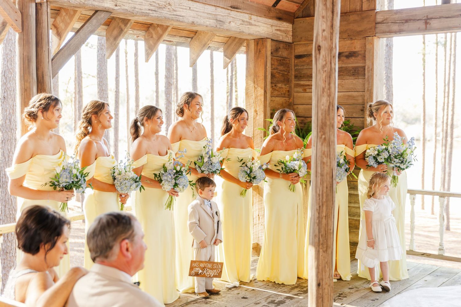 Bridesmaids in yellow dresses holding bouquets lined up at rustic barn wedding ceremony with flower girl.