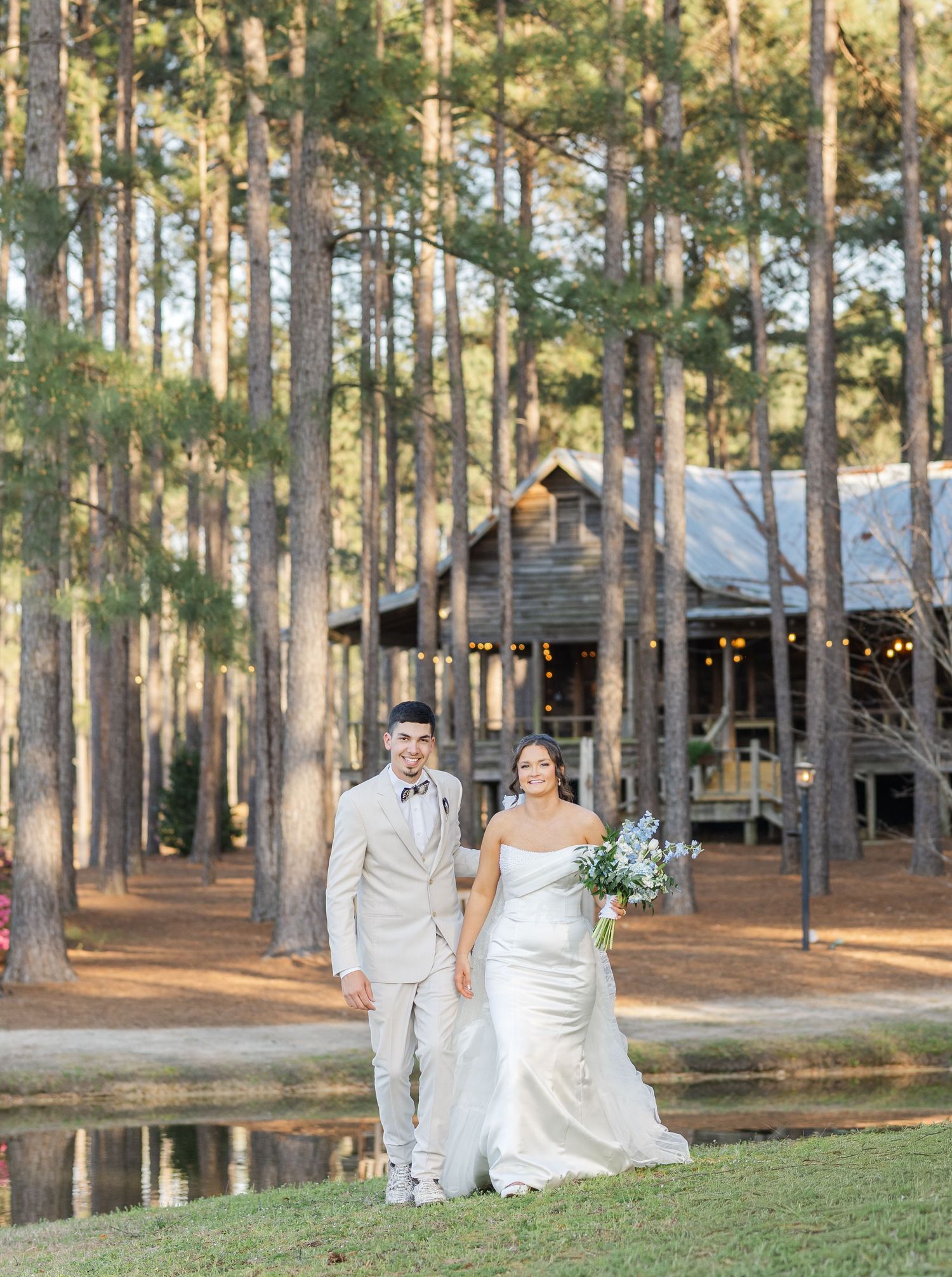 Bride holding blue and white bouquet and groom in tan suit stand before a rustic log cabin venue beside a pond.