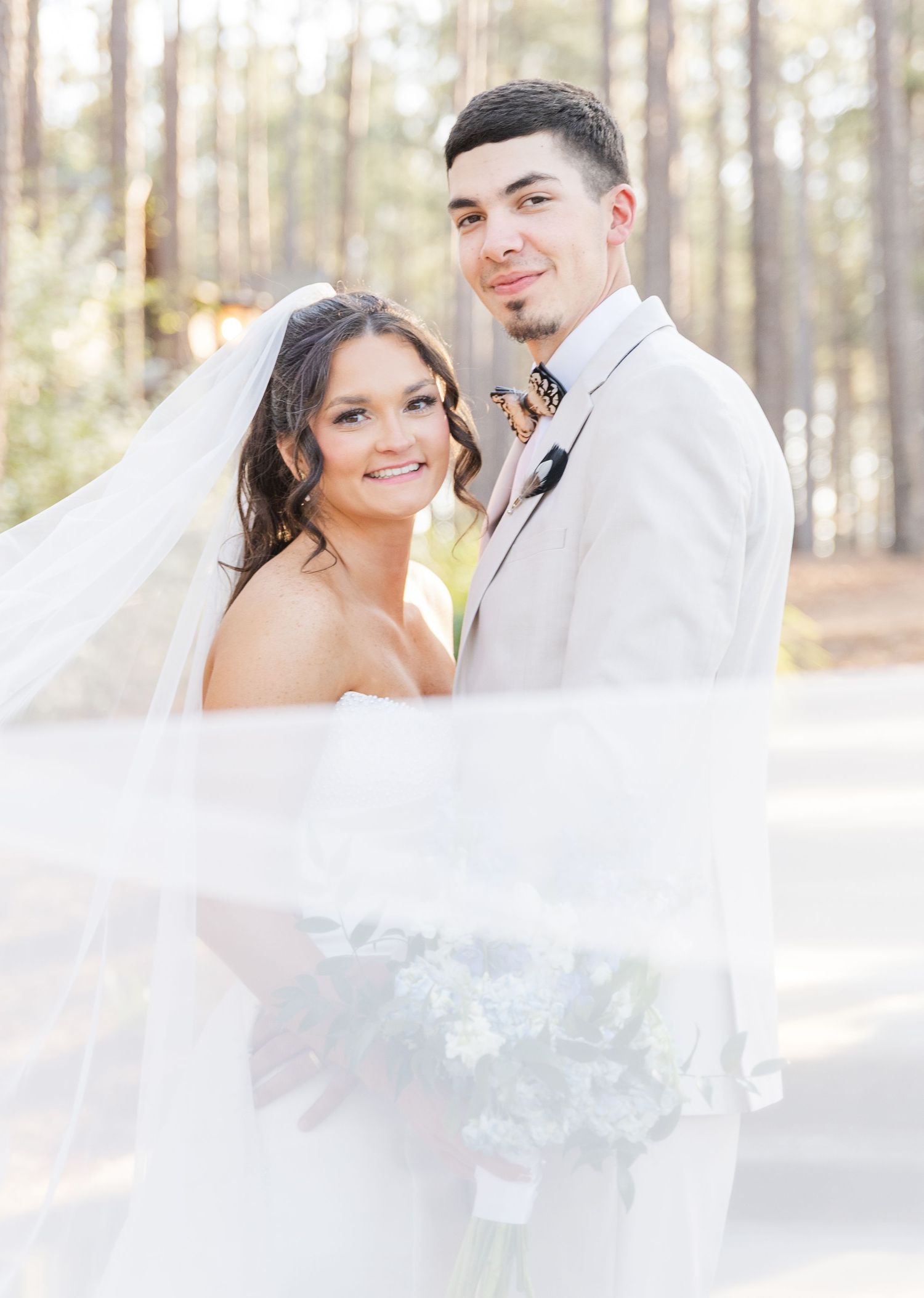 Bride and groom portrait with flowing veil in a sunlit pine forest, bride holding a blue bouquet.