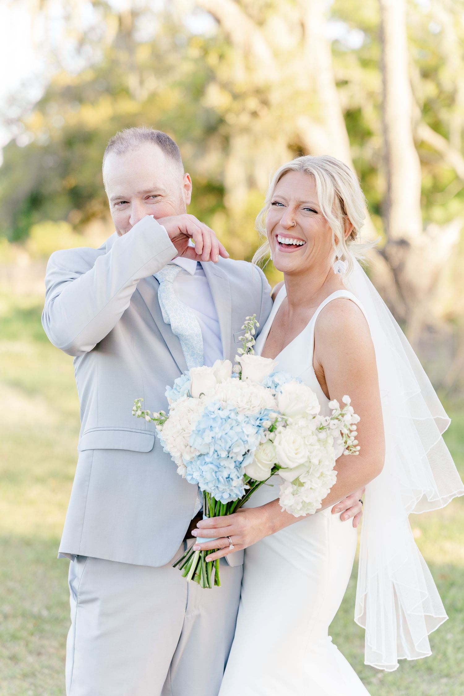 Joyful bride in white gown with blue hydrangea bouquet laughing beside emotional groom in gray suit.