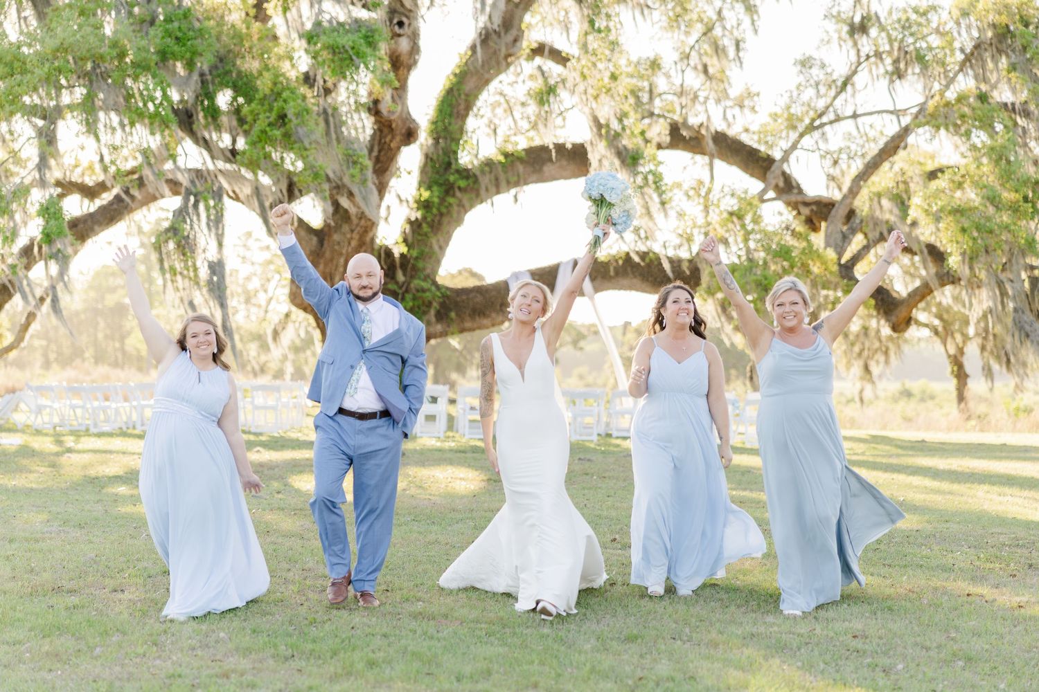 Bride, groom, and bridesmaids in blue dresses celebrating joyfully under large oak tree outdoors.