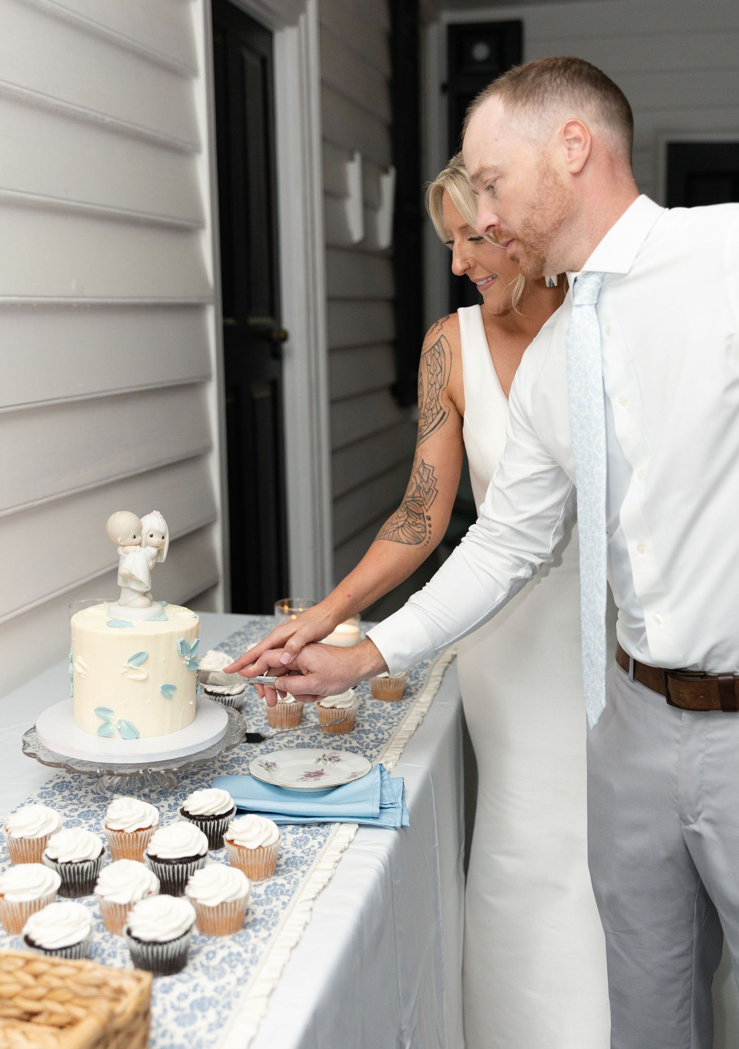 Bride and groom cut a white wedding cake decorated with blue accents surrounded by cupcakes.