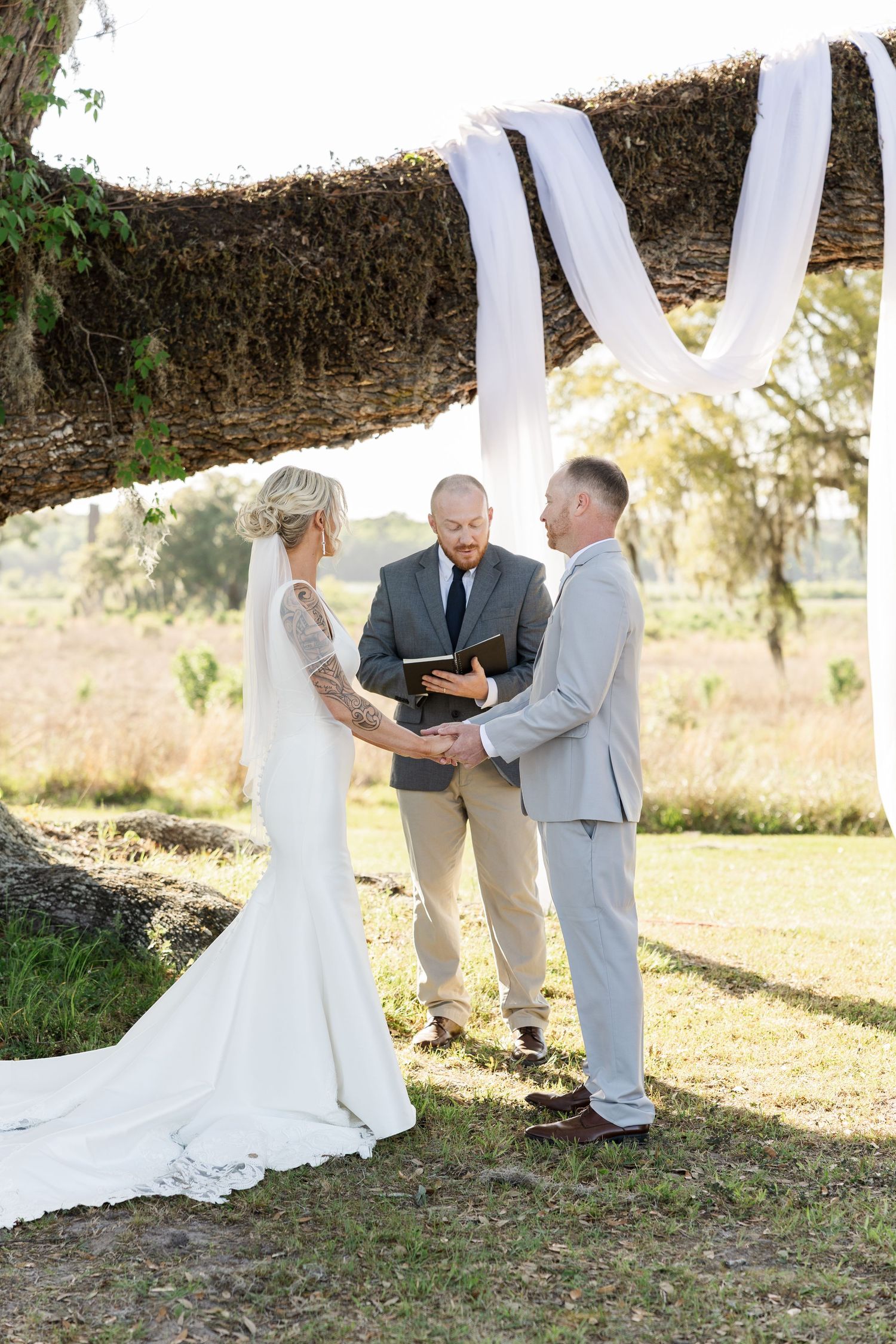 Bride and groom exchange vows with officiant under draped oak tree branch at outdoor wedding ceremony.