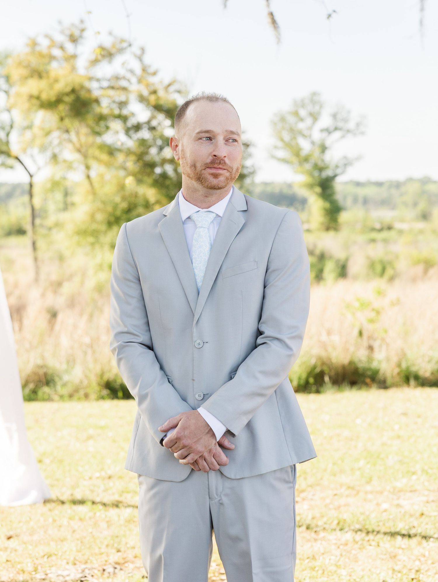 Groom in light gray suit standing at outdoor wedding ceremony altar surrounded by natural scenery.