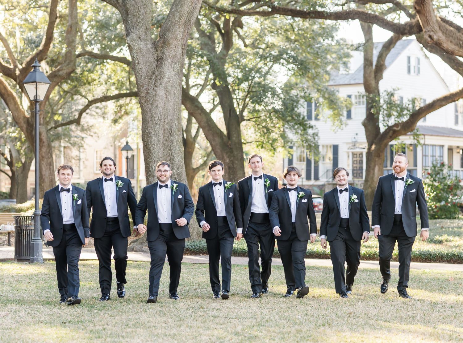 Groomsmen in classic black tuxedos stand in formation under oak trees for a formal wedding photo.
