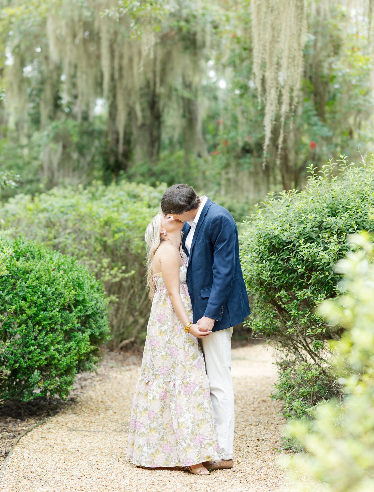 A romantic couple shares a tender moment on a tree-lined garden path with Spanish moss overhead.