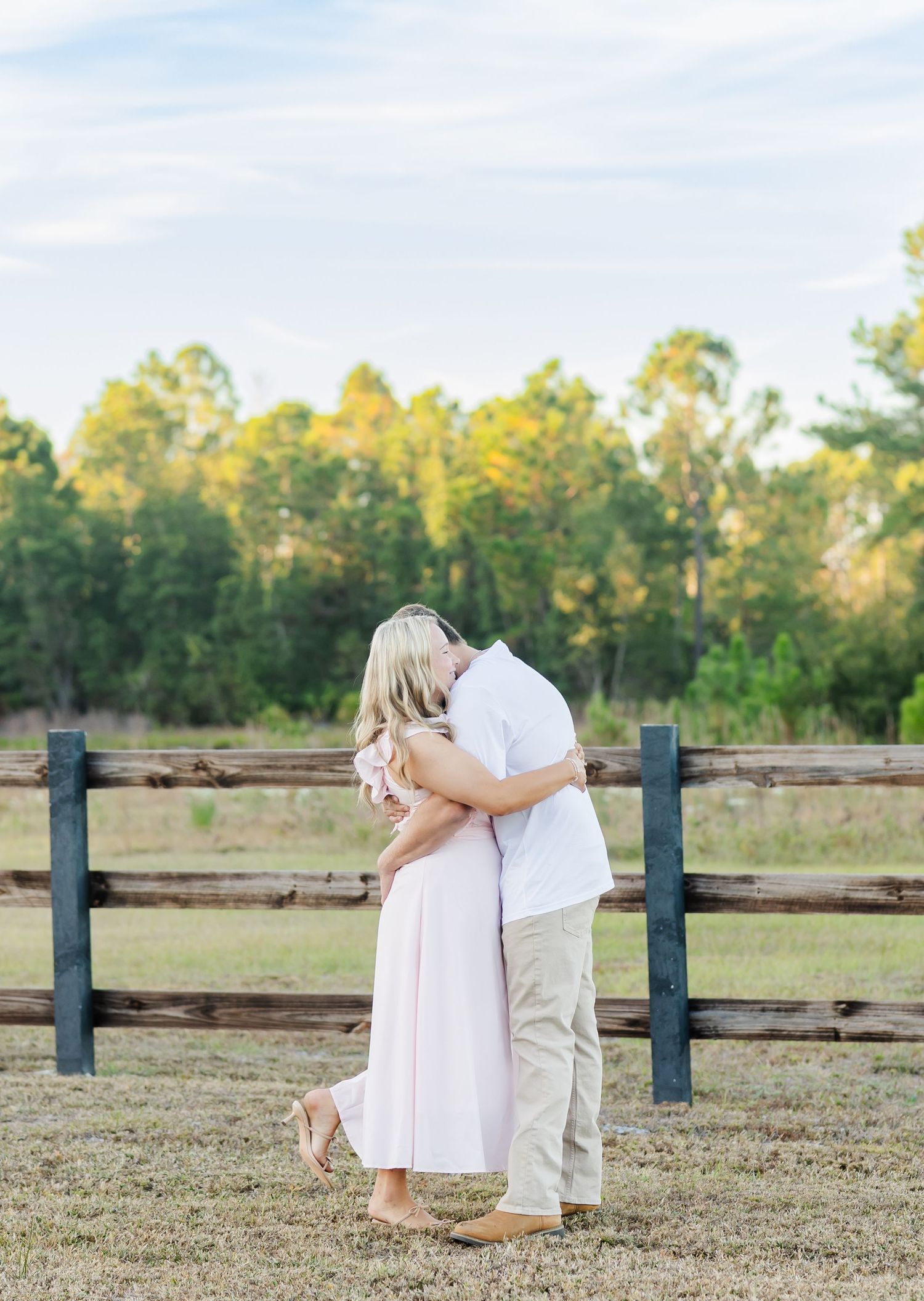 A tender moment between a couple in light colored outfits by a wooden fence at sunset.
