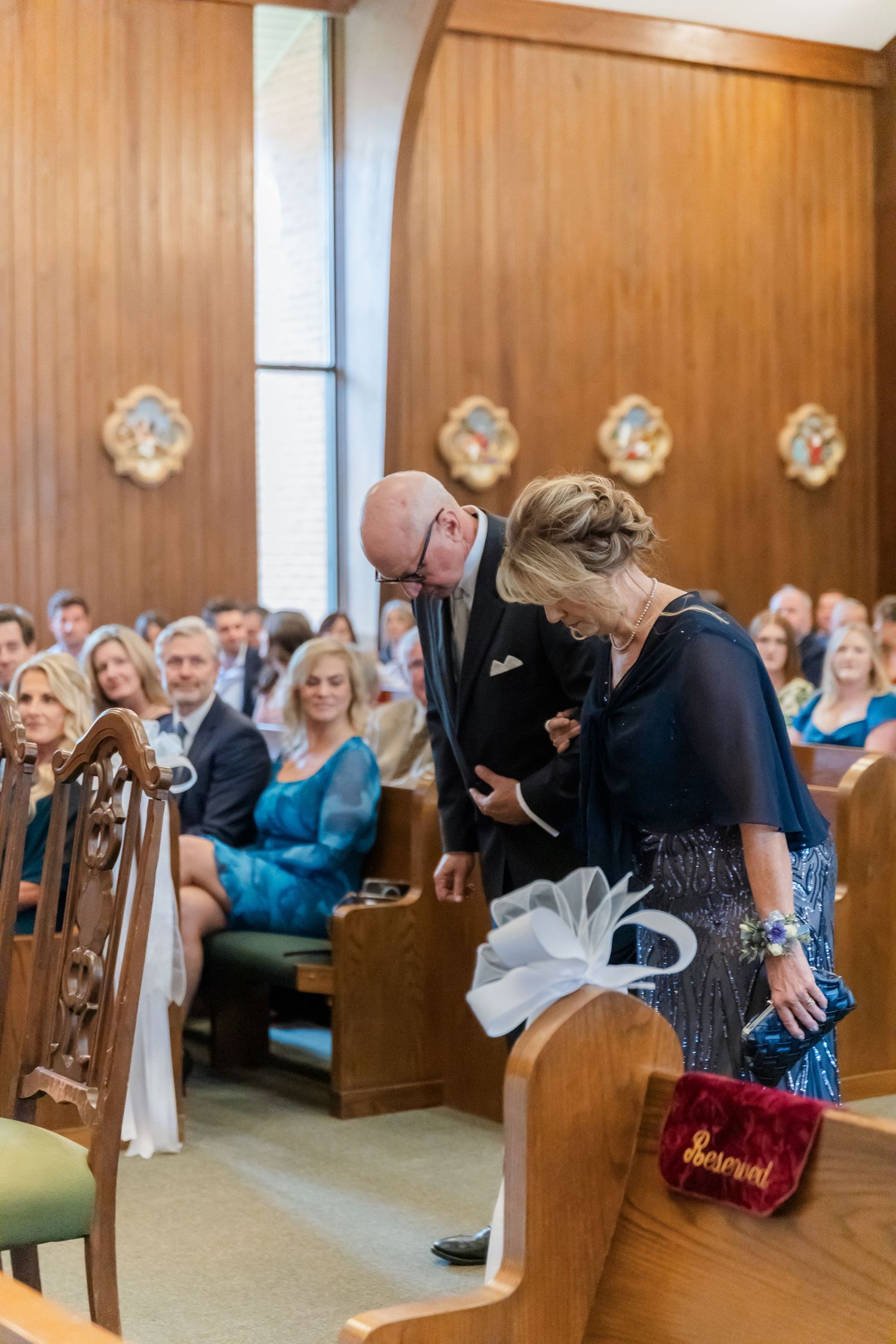Wedding guests seated in wooden pews inside a church with wood paneled walls and floral decorations.