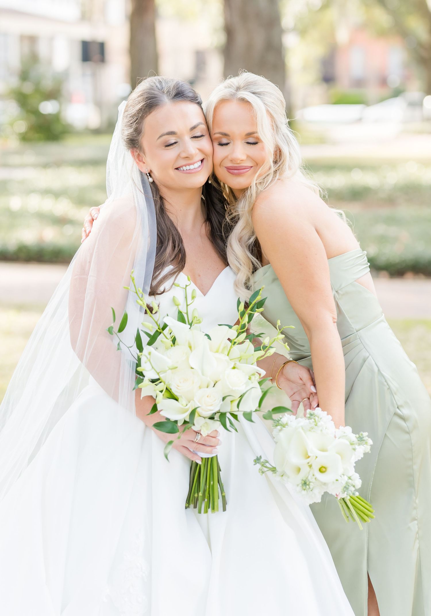 Two attendants in pastel dresses share a joyful moment while holding white floral bouquets in an outdoor setting.