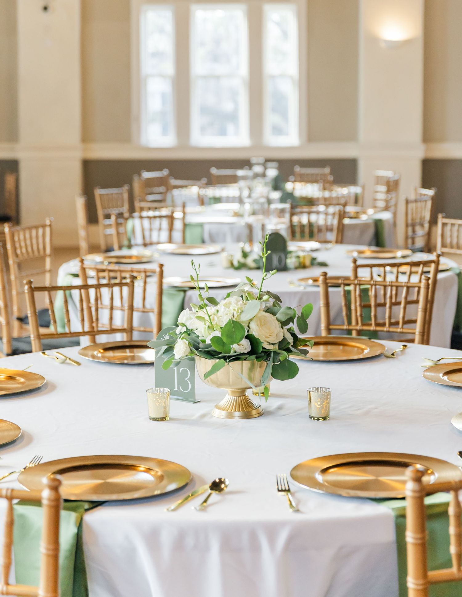 Wedding reception venue with gold chiavari chairs and white tablecloths decorated in sage green and gold accents.
