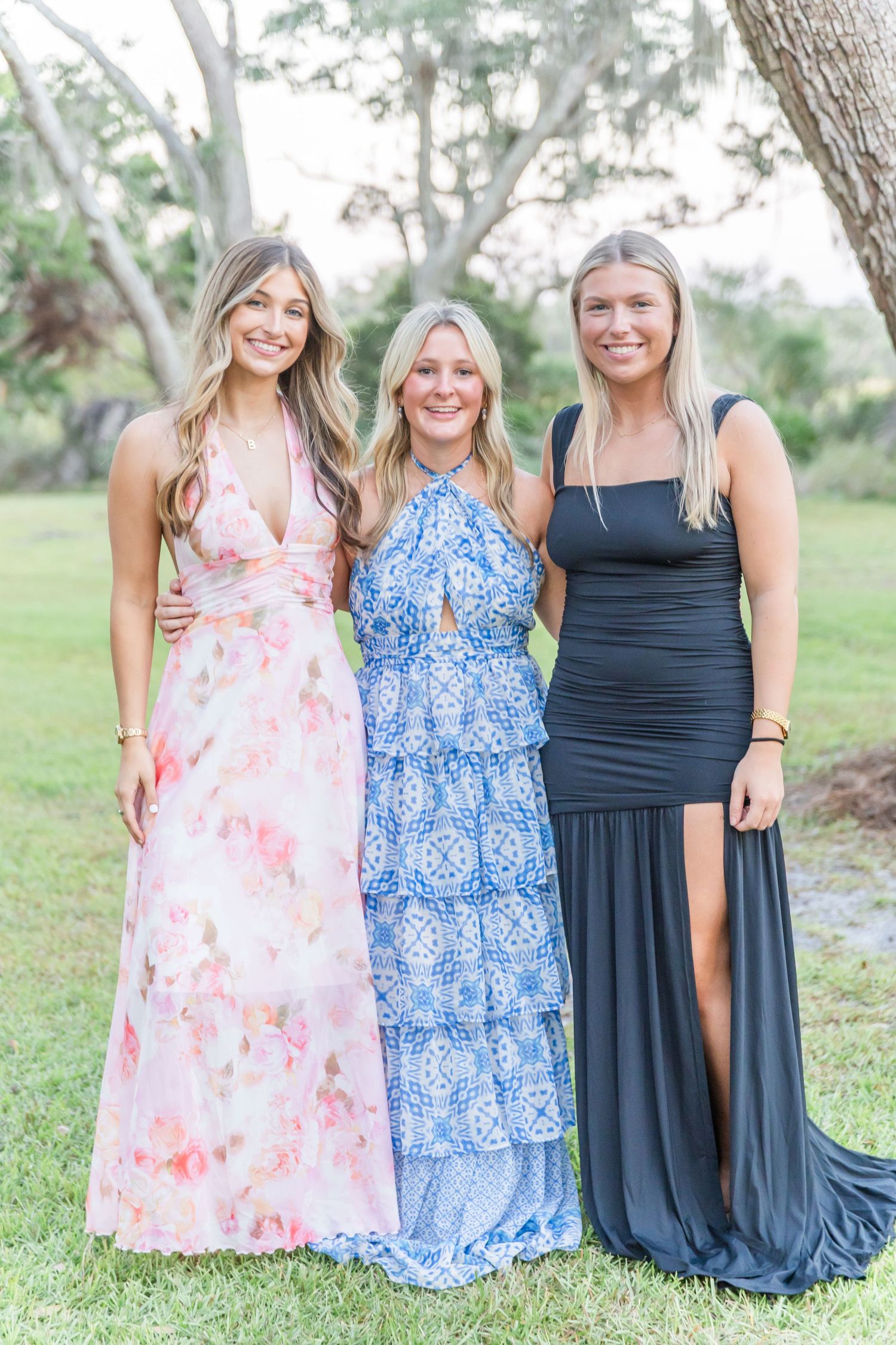 Three women pose together outdoors wearing floral pink, blue patterned, and navy dresses.