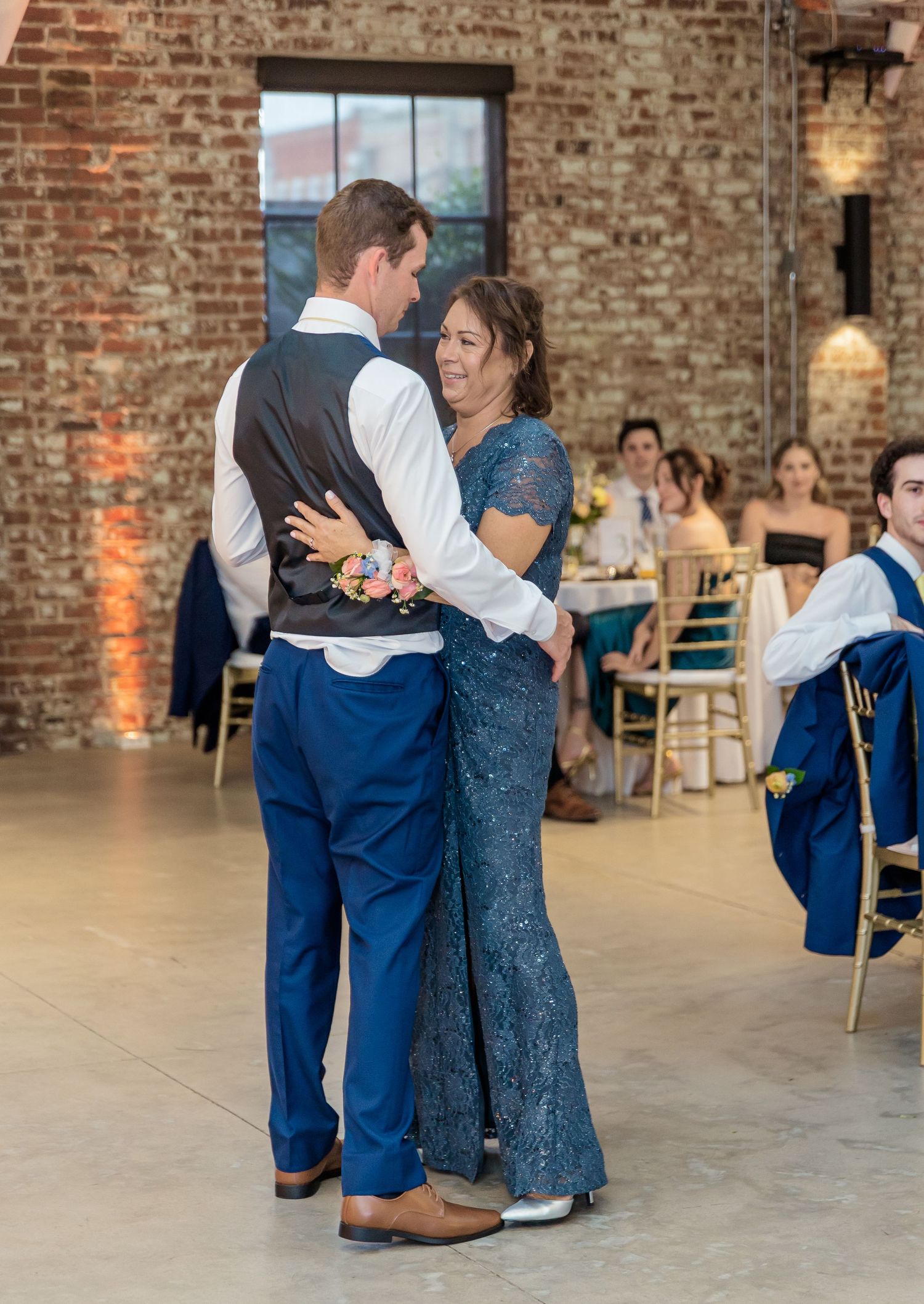 A couple dances together at a wedding reception in an industrial brick venue while guests look on from their tables.