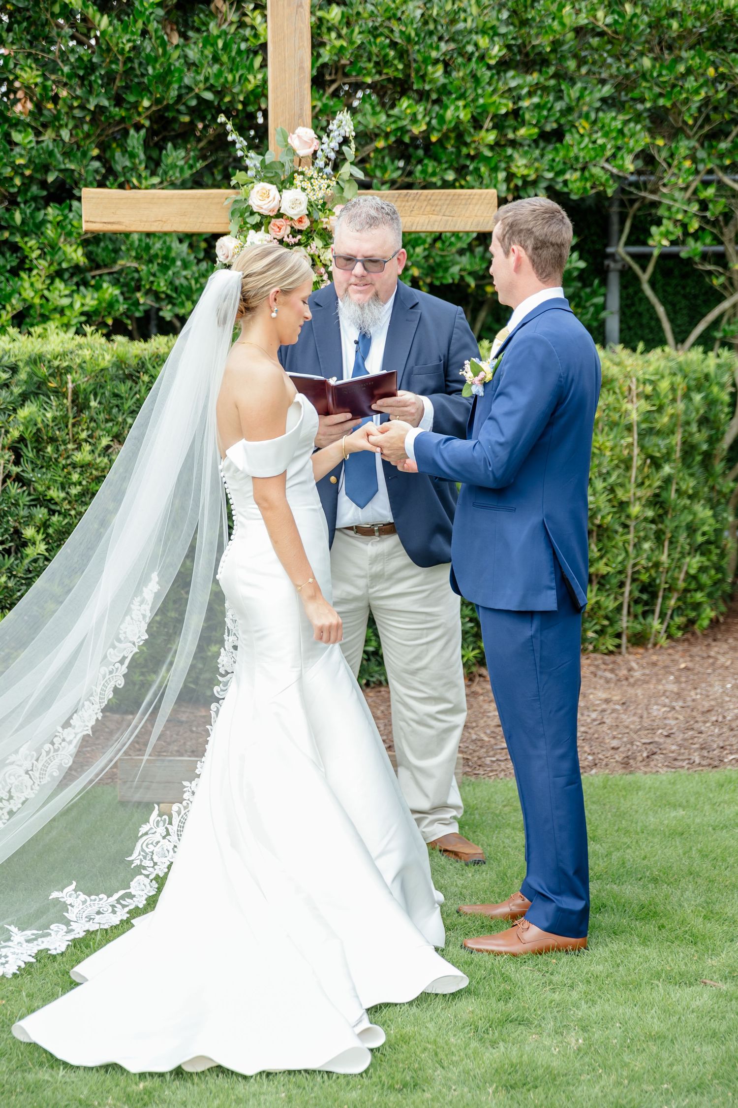 Wedding ceremony taking place outdoors with couple exchanging rings on green lawn surrounded by garden foliage.