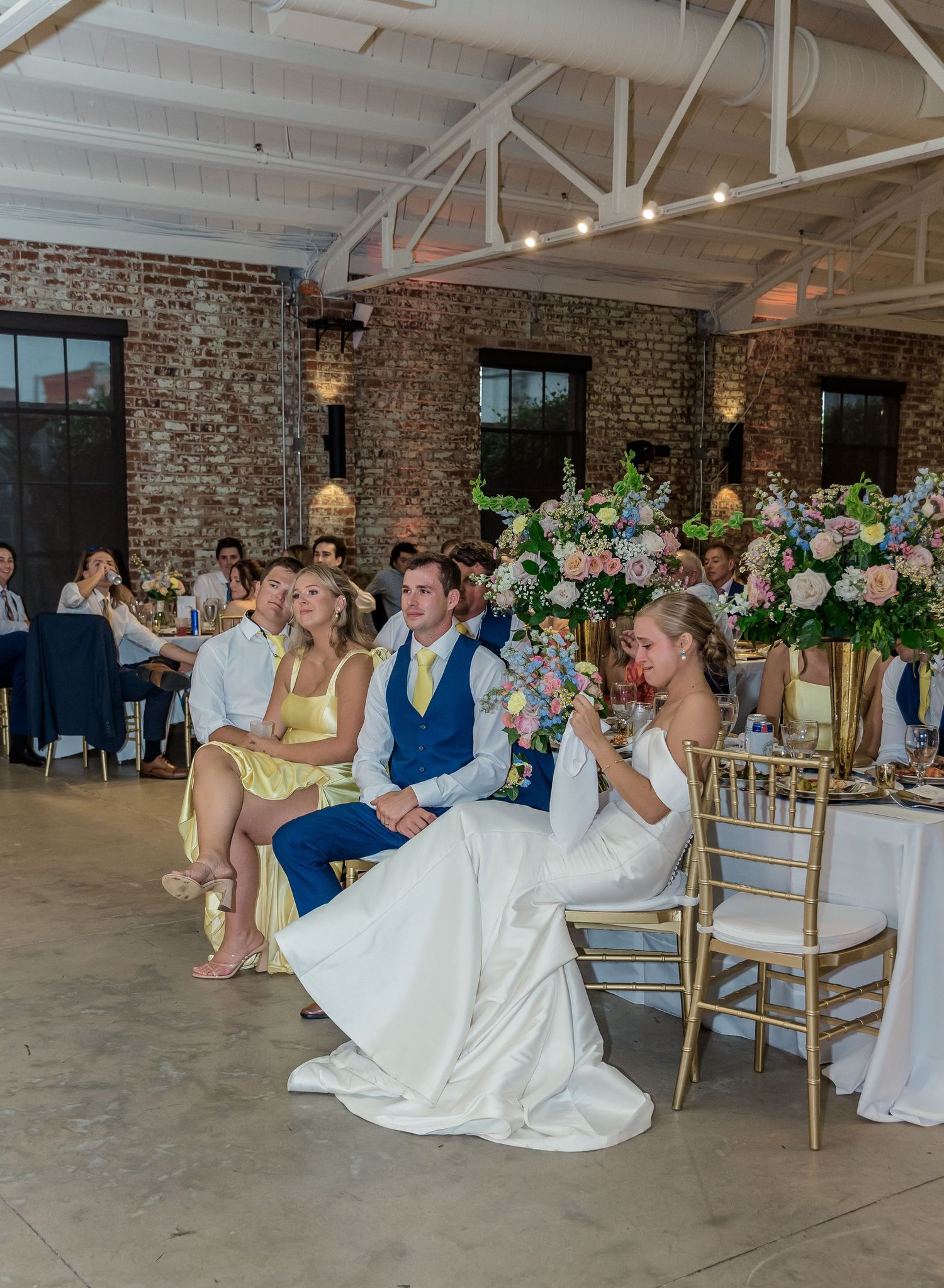 Wedding reception in a rustic brick venue with guests seated at decorated tables with floral centerpieces.
