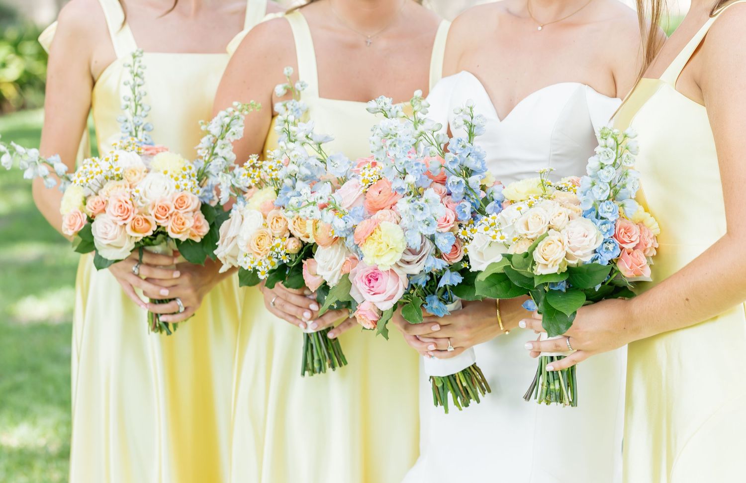 Row of bridesmaids in pale yellow dresses holding pastel bouquets with pink and blue hydrangeas at outdoor wedding.