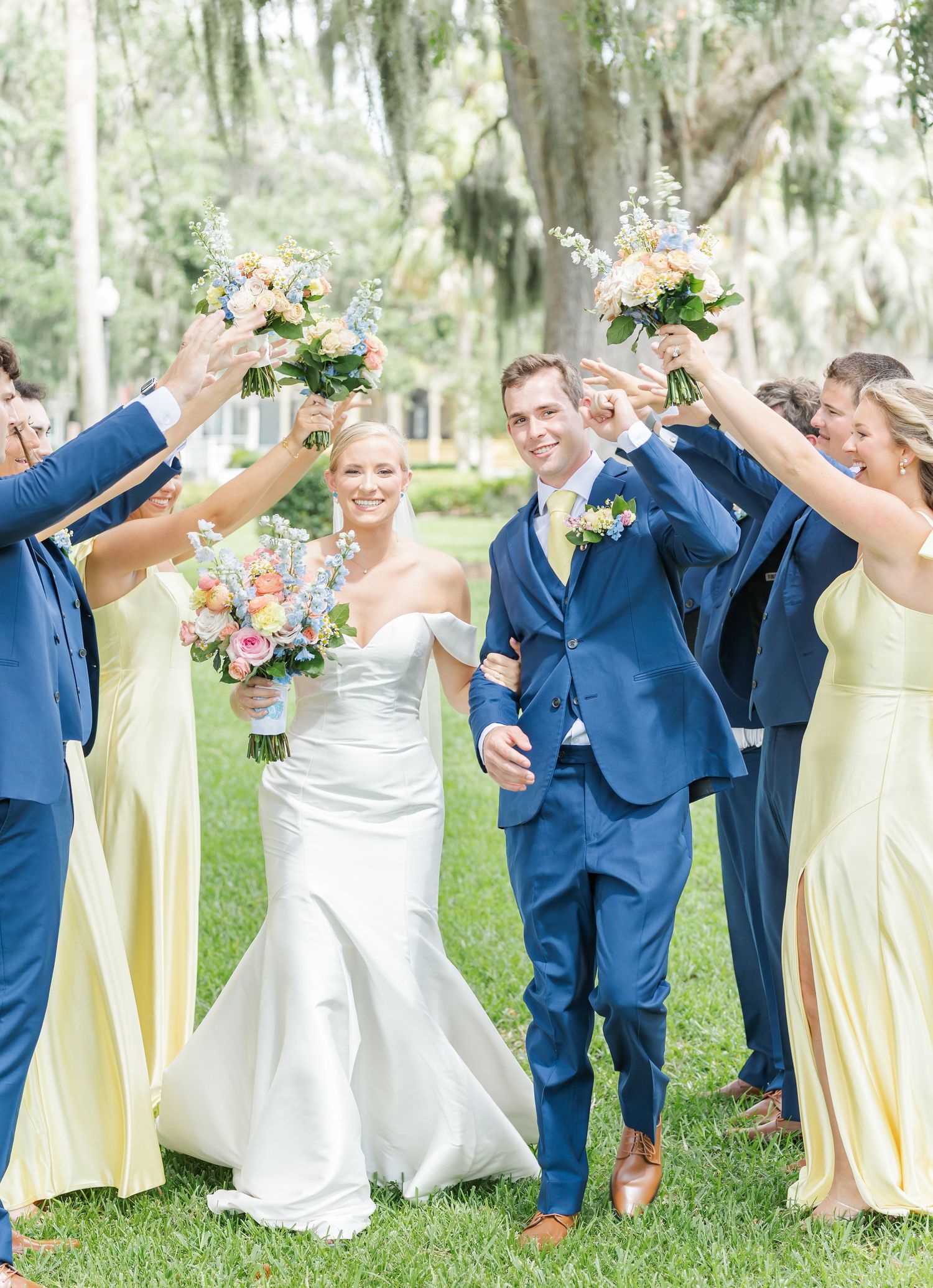 Wedding party in navy suits and pale yellow dresses poses playfully in an outdoor garden setting.
