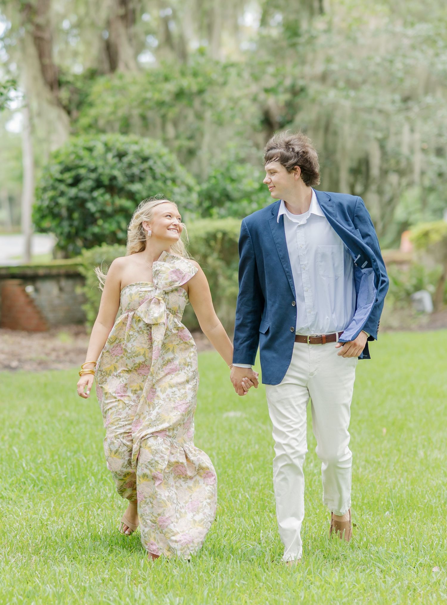 A couple runs playfully through a green park with Spanish moss hanging from trees on a sunny day.
