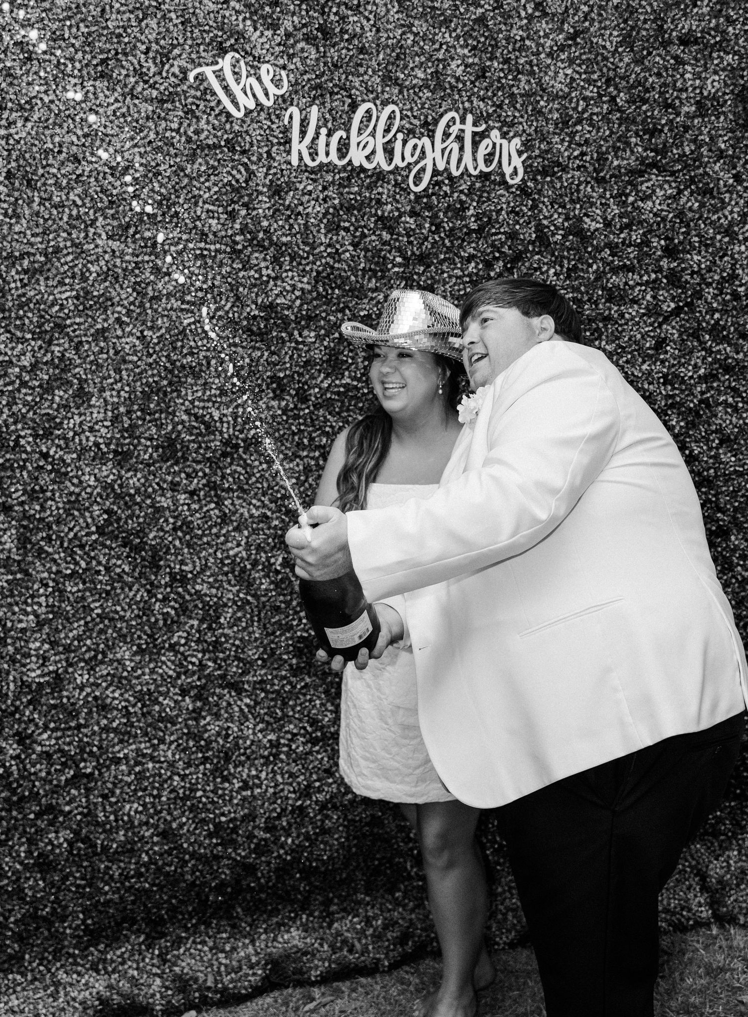 Newlyweds pop champagne in black and white photo at The Ricklighters wedding photo booth backdrop.