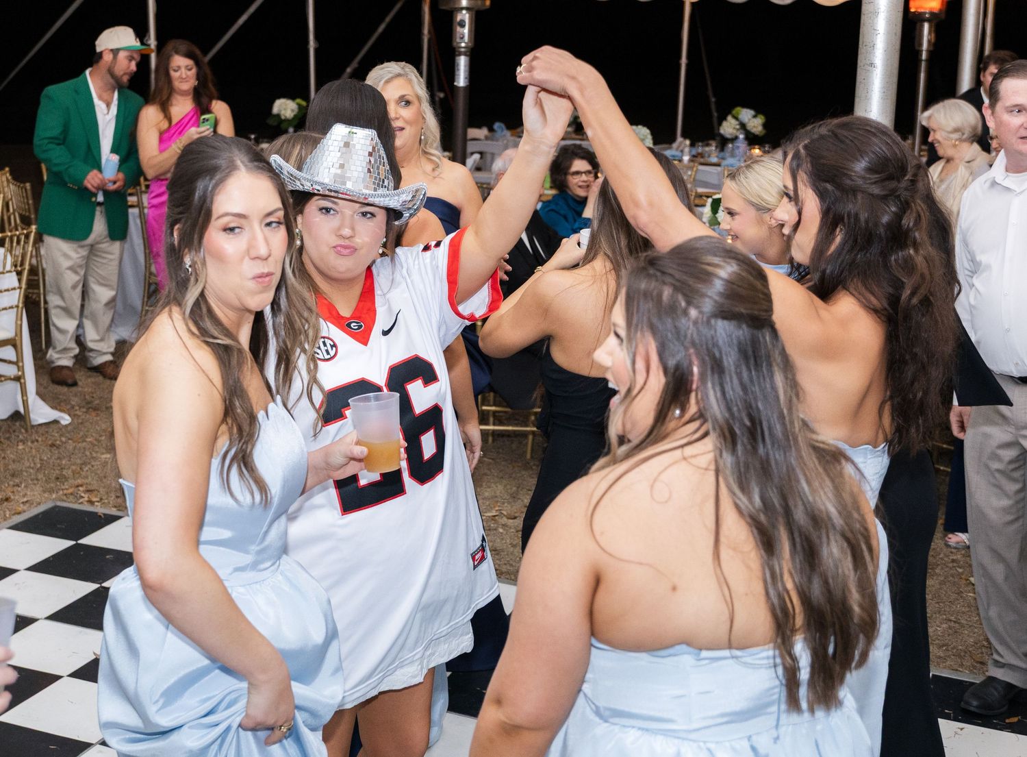 Bridesmaids in blue dresses dancing with guest wearing football jersey and silver cowboy hat at wedding reception.