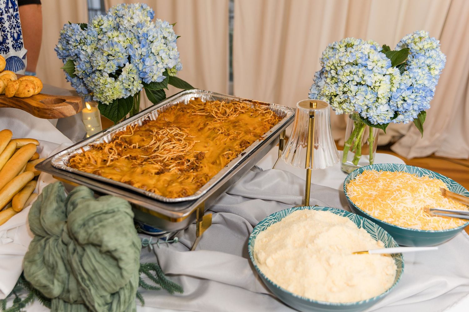 Wedding reception buffet with pasta bake, mac and cheese, and blue hydrangea floral centerpieces on white tablecloth.